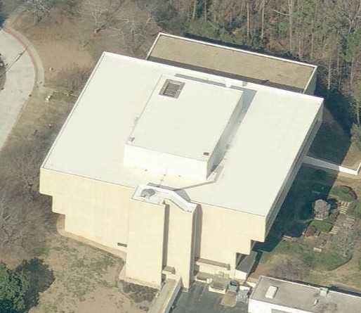Aerial view of a large, beige, rectangular building with a flat white roof, surrounded by trees and pavement—a perfect project for a roofing contractor specializing in commercial roof repair. Small parking area and outdoor seating are visible nearby.