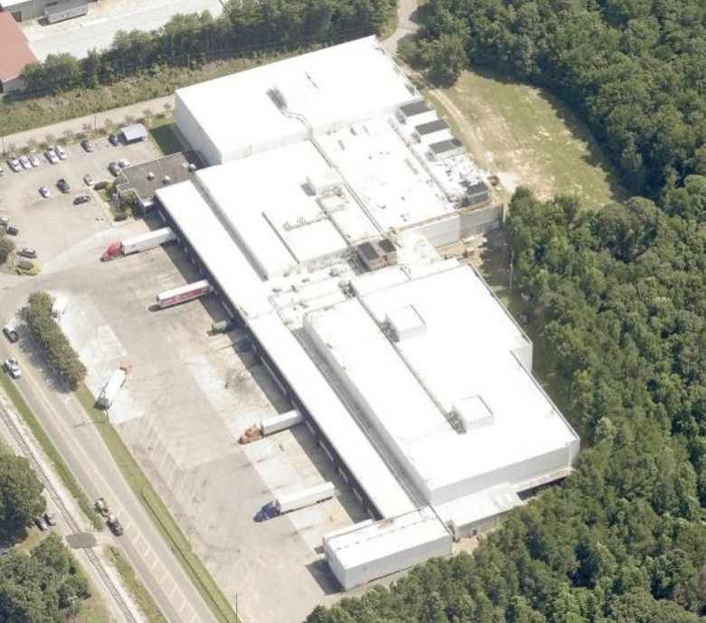 Aerial view of a large, white industrial building with an adjacent parking lot, surrounded by trees and greenery, with a commercial roofer working on the roof and a road running along one side of the property.
