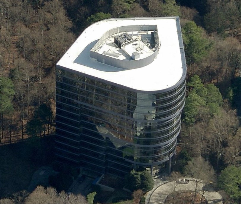Aerial view of a modern, glass-covered office building with a white, uniquely shaped roof—an ideal project for a skilled roofing contractor—surrounded by leafless trees and greenery.