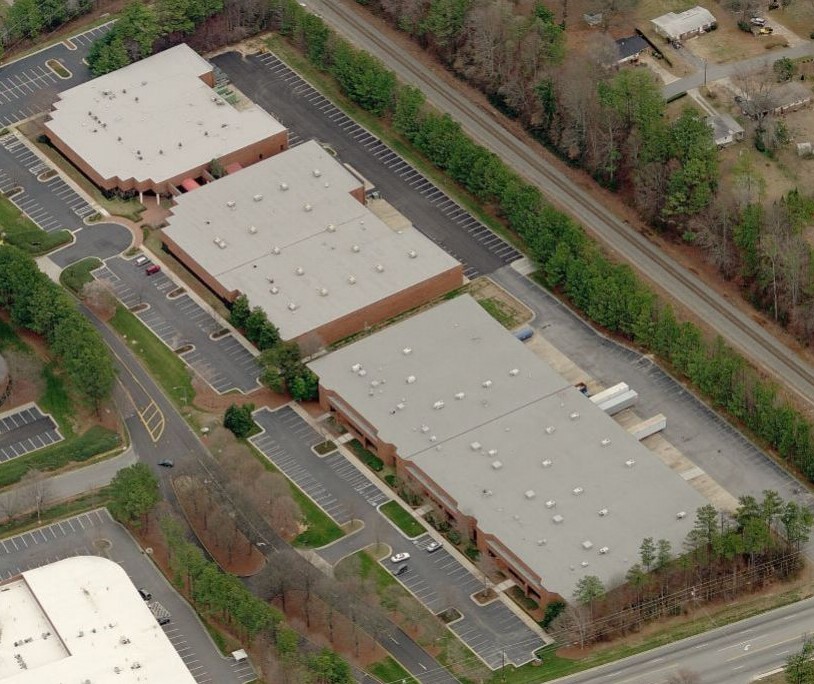 Aerial view of several large, flat-roofed industrial or office buildings featuring commercial roofing, with surrounding parking lots bordered by trees and roads. Few cars are visible in the parking areas.