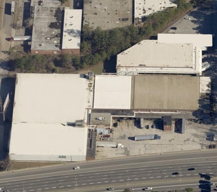 Aerial view of an industrial complex with large white-roofed buildings, a parking area with trucks, nearby trees, and a busy multi-lane road—ideal for showcasing the scale of commercial roof repair by an expert commercial roofer.