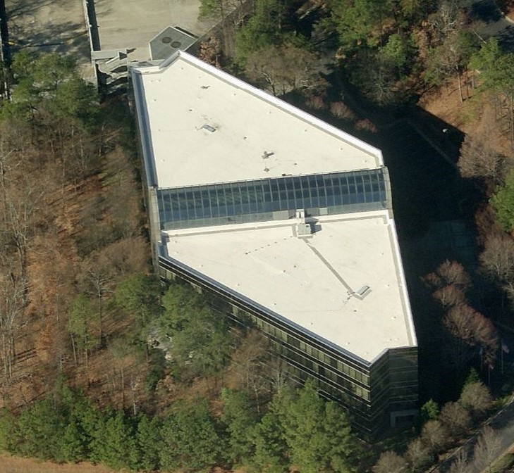 Aerial view of a unique, triangular-shaped building with a white roof, featuring a central glass section along its length—an impressive example of commercial roofing nestled among trees.