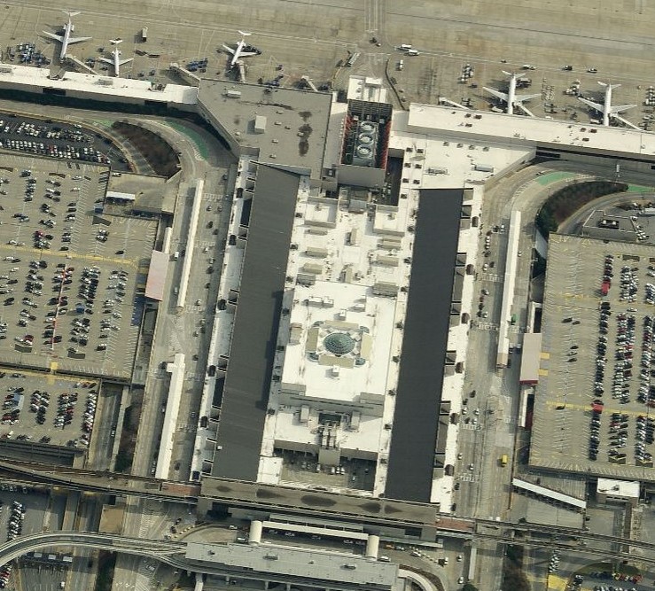 Aerial view of an airport terminal with multiple airplanes parked at gates, its expansive roofs maintained by skilled commercial roofers, surrounded by parking lots full of cars and various service roads.