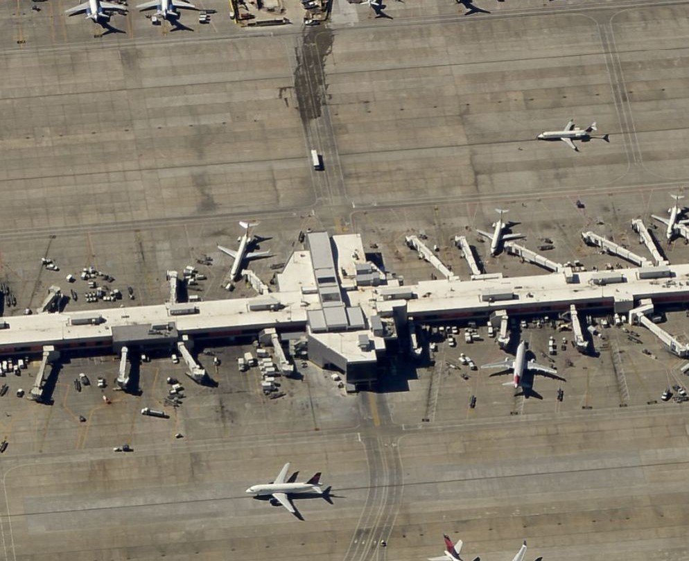 Aerial view of an airport terminal with multiple airplanes parked at gates, various ground vehicles, and service equipment scattered across the tarmac. The expansive commercial roofing of the terminal stands out amid the bustling aviation activity.