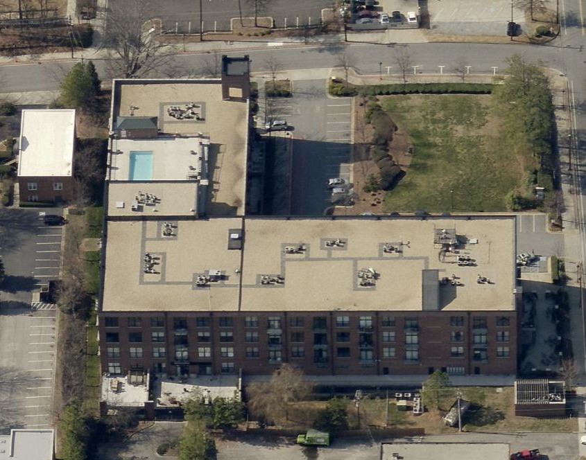 Aerial view of a rectangular, flat-roofed building with a small pool, adjacent parking lots, grassy area, and surrounding trees. This multi-story structure in an urban setting may benefit from the expertise of a commercial roofer.