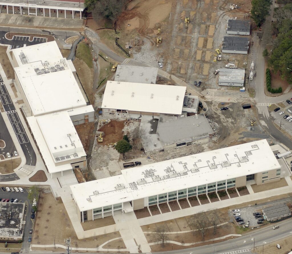 Aerial view of a construction site with several large white-roofed buildings, where a commercial roofing contractor and construction vehicles are at work amid exposed soil, surrounded by trees and roads.