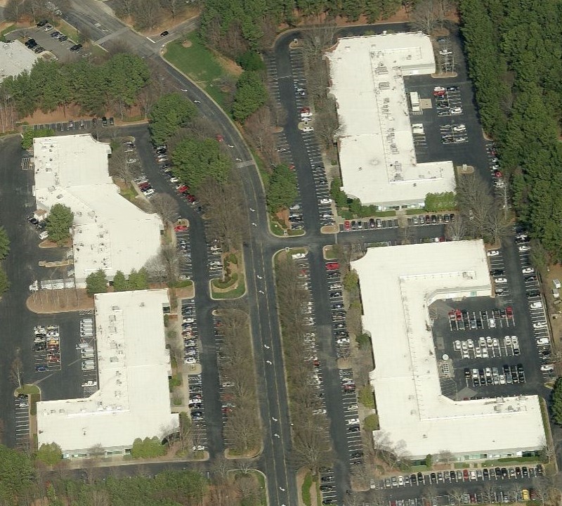 Aerial view of three large office buildings with white roofs, possibly maintained by a commercial roof repair contractor, surrounded by parking lots filled with cars, bordered by trees and roads.