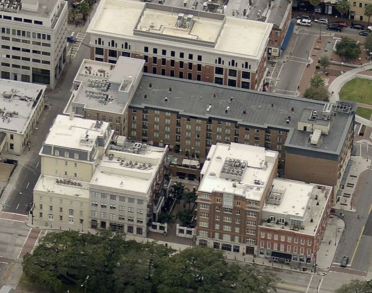 Aerial view of several multistory buildings arranged in a block along city streets, featuring white and gray commercial roofing, trees in the foreground, and a mix of modern and historic architecture.