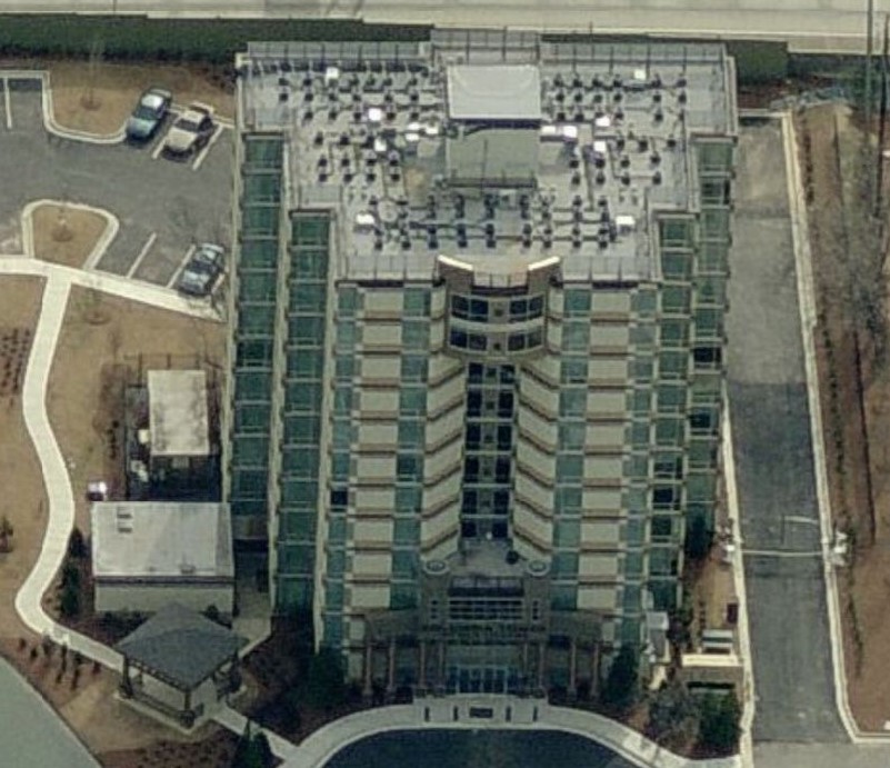 Aerial view of a multi-story building with a modern glass facade, rooftop equipment installed by a commercial roofer, and a circular driveway entrance. Parking lots and landscaped areas with paths surround the building.