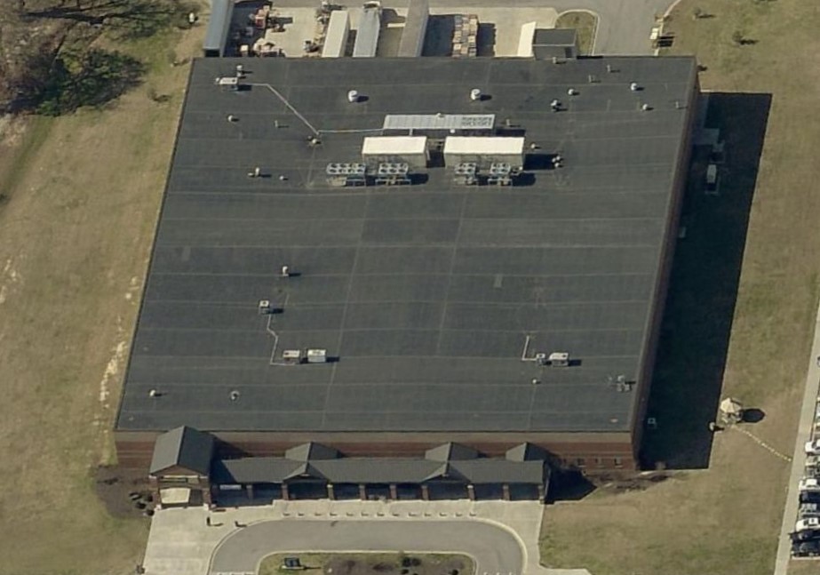 Aerial view of a large rectangular industrial or commercial building with a flat black roof installed by a commercial roofer, rooftop equipment, and a parking lot with a few cars. The building has multiple entrances at the front and is surrounded by grass.