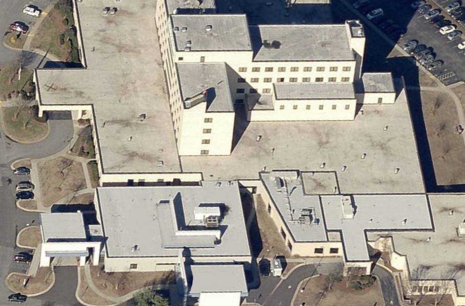 Aerial view of a large, multi-story hospital complex with several connected buildings featuring commercial roofing, white rooftops, parking areas, and surrounding roads. Cars are parked along the edges of the facility.