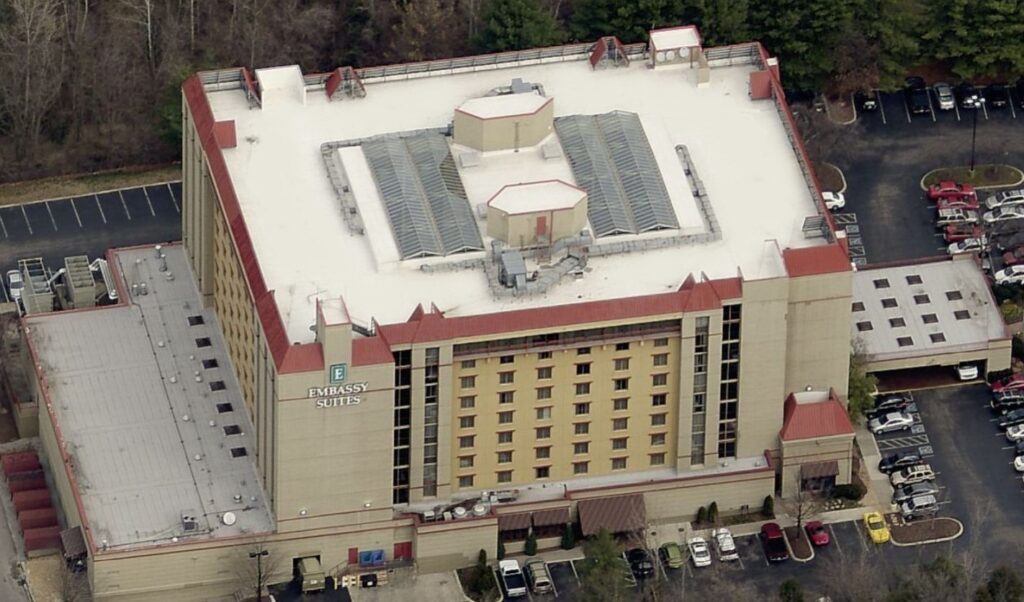 Aerial view of an Embassy Suites hotel with a white flat roof, beige exterior walls, red roof accents, and multiple windows—perfect for showcasing commercial roofing expertise—surrounded by parked cars and bordered by trees.