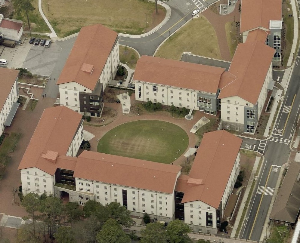 Aerial view of a large building complex with red-tiled roofs arranged around a central grassy courtyard, showcasing the impressive work of a commercial roofer. The buildings have multiple floors, connected by walkways and surrounded by roads and trees.