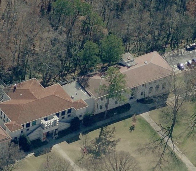 Aerial view of a large, light-colored building with a red-tiled roof, surrounded by trees and grassy areas. Several cars are parked along a road beside the building—ideal for a commercial roofing contractor project.