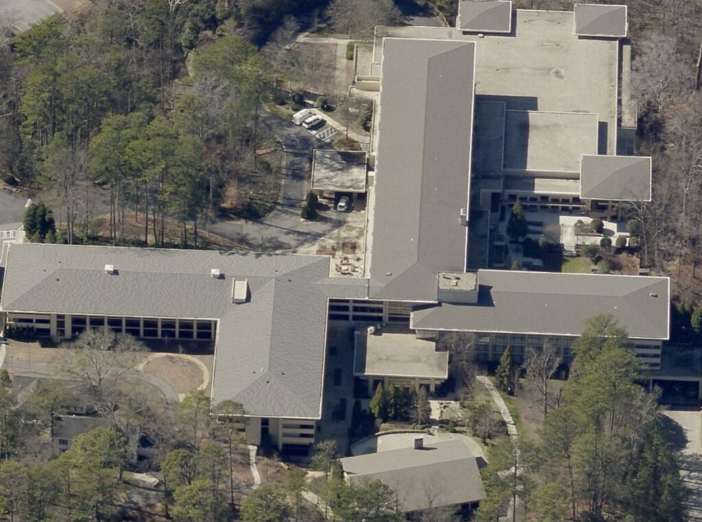 Aerial view of a large, modern building complex with flat, gray rooftops—an impressive example of commercial roofing—surrounded by trees and parking areas, featuring multiple connected sections.