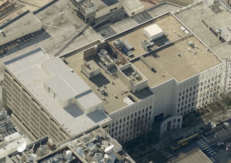 Aerial view of a large, rectangular, flat-roofed office building with HVAC units on the roof—ideal for a roofer specializing in commercial roof repair—surrounded by cityscape, parking lots, and streets with cars and trees.