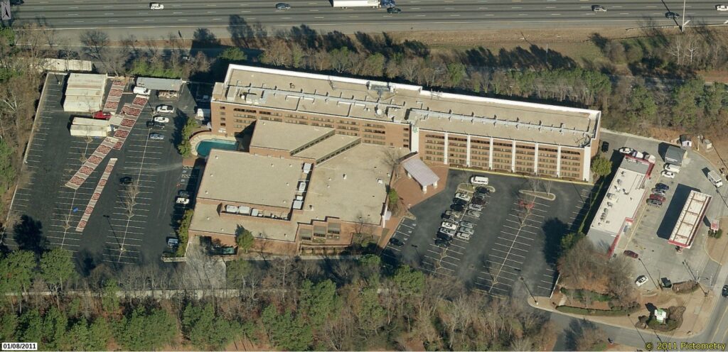 Aerial view of a large hotel complex with parking lots, outdoor pool, and nearby roadways, bordered by trees. The expansive roof showcases the work of a skilled commercial roofing contractor. A major highway runs along the top edge of the image.