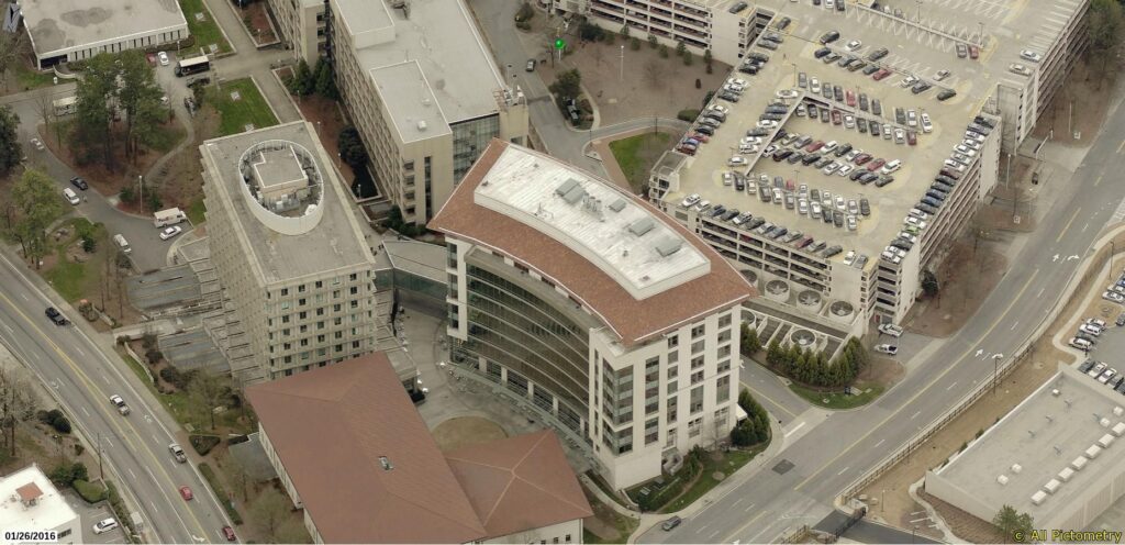 Aerial view of a curved office building with a red roof, surrounded by other buildings, roads, and a large parking lot filled with cars. Trees and greenery are scattered throughout the area, highlighting the need for expert commercial roofing services.