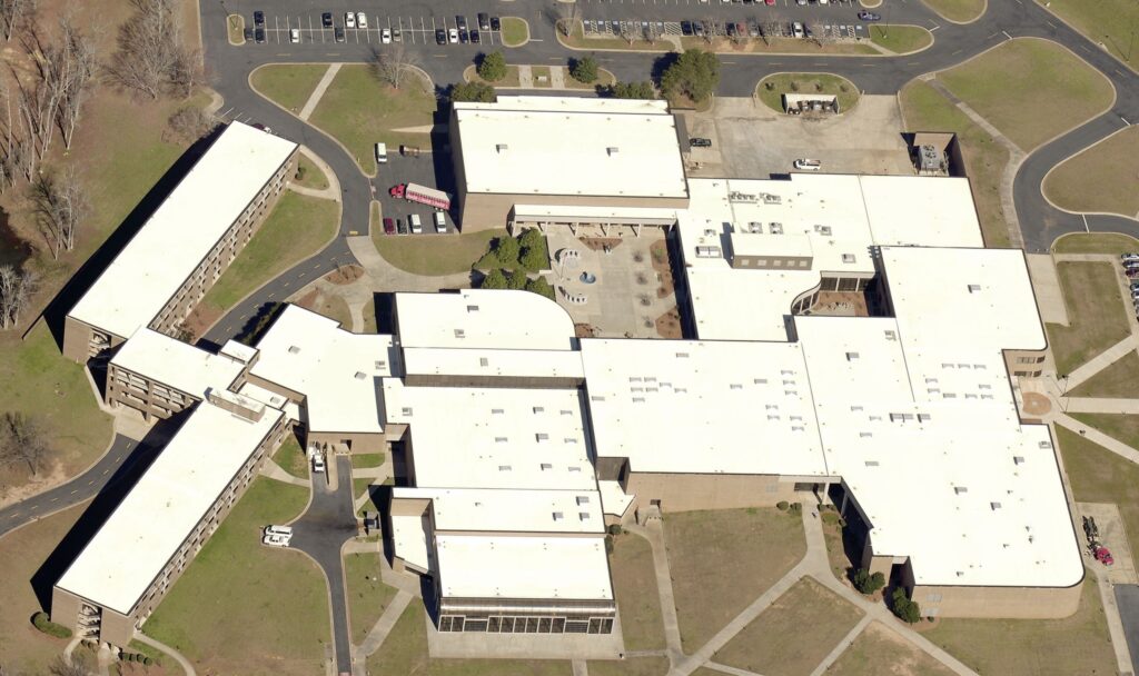 Aerial view of a large school or institutional building with multiple connected wings, showcasing its expansive commercial roofing, surrounded by parking lots, driveways, grassy areas, and a few scattered trees.