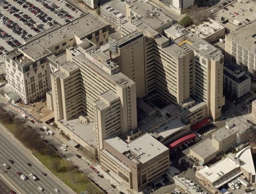 Aerial view of a large hospital complex with commercial roofing across multiple connected buildings, a rooftop helipad, nearby parking lots filled with cars, and adjacent roads with moving traffic.