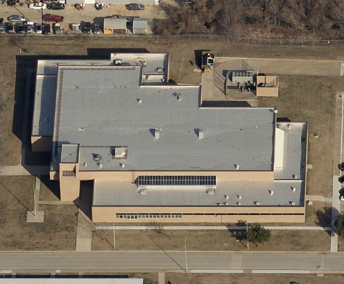 Aerial view of a large, light gray-roofed building with multiple sections, expertly maintained by a commercial roofer, featuring parking areas and green space, surrounded by roads and some vehicles parked nearby.