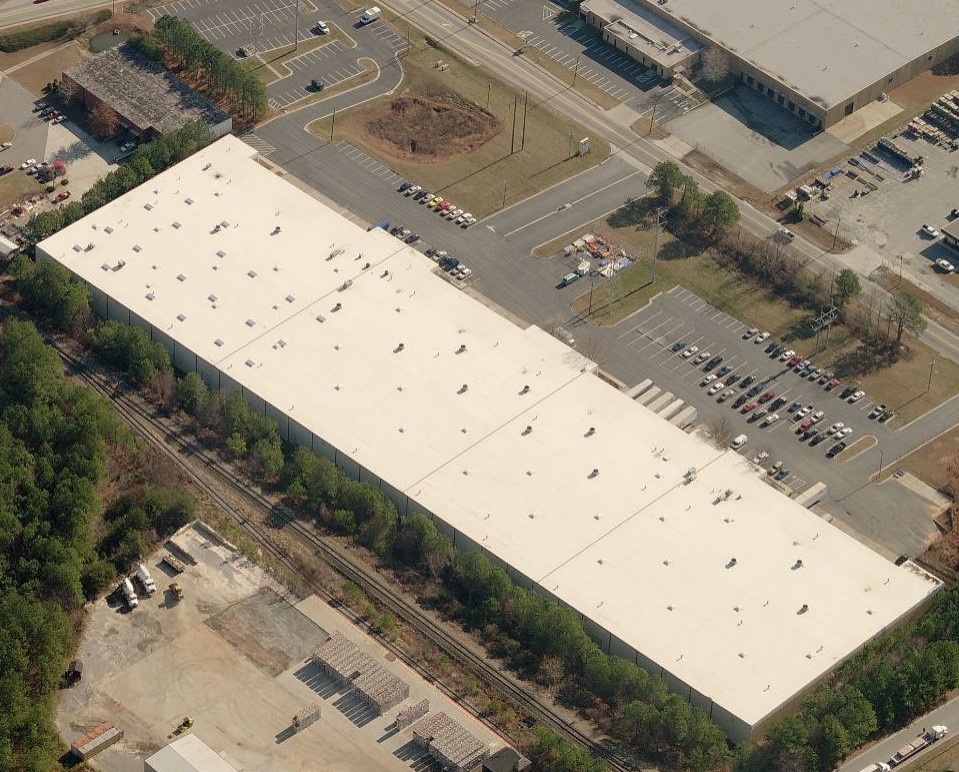Aerial view of a large warehouse with a white roof, surrounded by parking lots filled with cars and trucks. The building is near a road, with trees and other industrial buildings nearby—a great example for commercial roof repair by an expert roofing contractor.