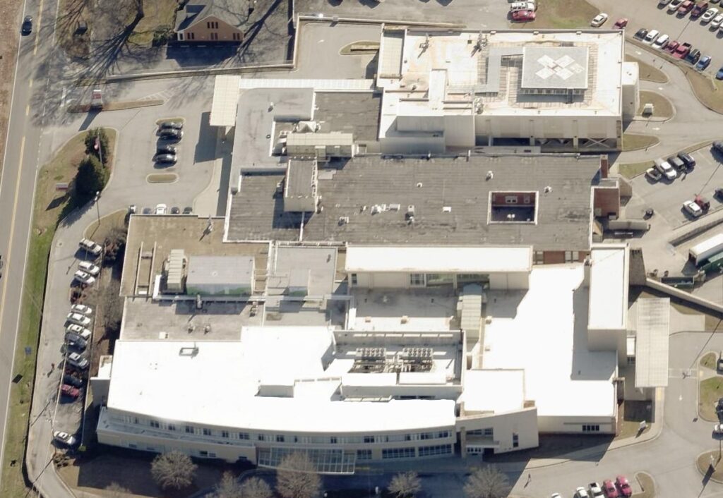 Aerial view of a large, multi-winged hospital with white and gray commercial roofing, surrounded by parking lots, cars, and some trees. Roads encircle the complex.
