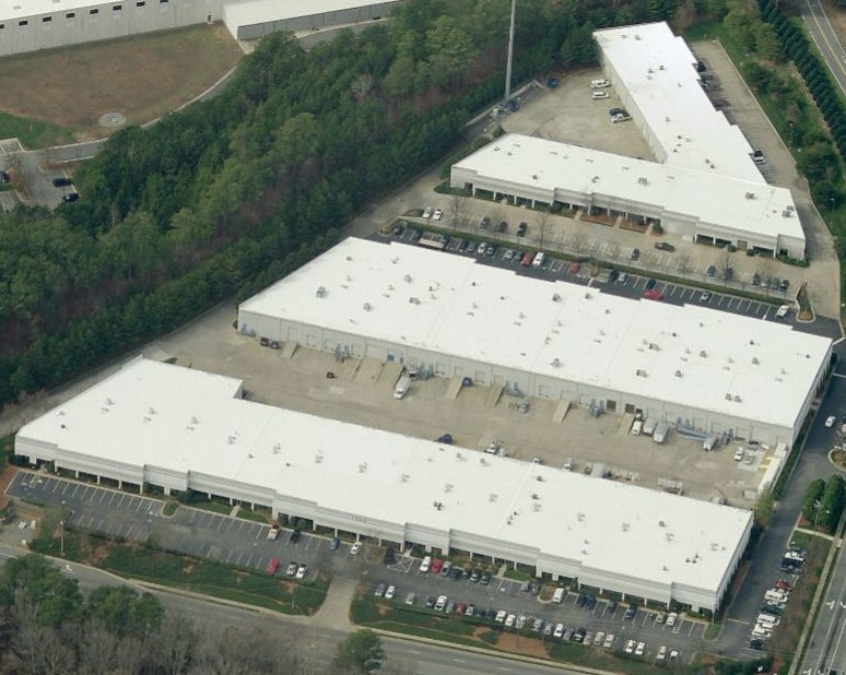 Aerial view of a large industrial warehouse complex with multiple connected commercial roofing structures, loading docks, parked trucks, cars, and surrounding trees.