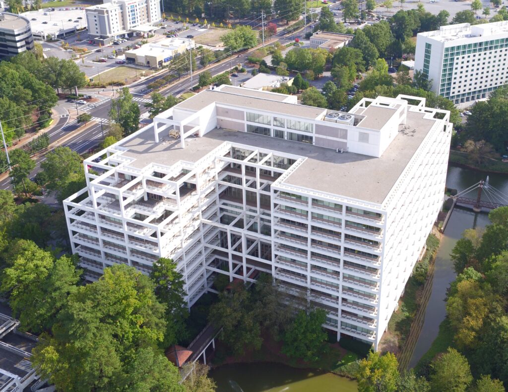 Aerial view of a large, modern white office building with a geometric design and commercial roofing, surrounded by trees and a small lake, with roads, cars, and other buildings visible in the background.