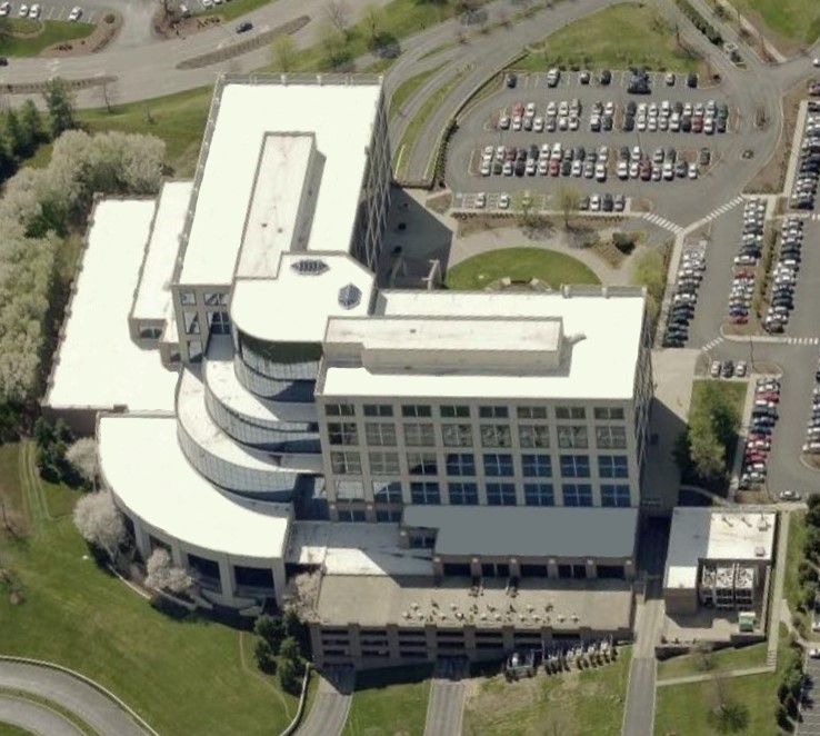 Aerial view of a large modern building with white commercial roofing, featuring curved and rectangular sections, surrounded by green lawns, trees, roads, and multiple parking lots filled with cars.