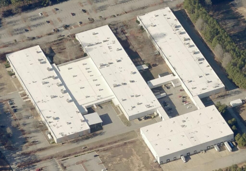 Aerial view of a large industrial building with a white roof, serviced by a commercial roofer, surrounded by parking lots and parked cars. Trees border the property, with patches of open space around the facility.
