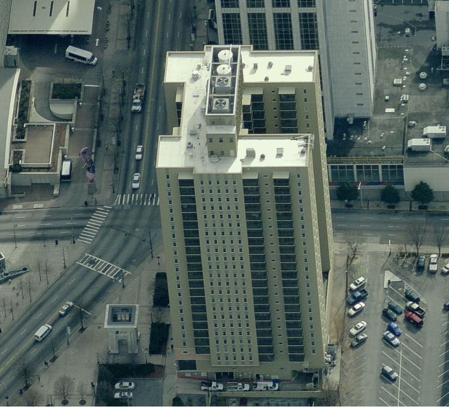 Aerial view of a tall, rectangular beige building with many windows, its commercial roofing standing out amid streets, parked cars, and nearby buildings in an urban area.