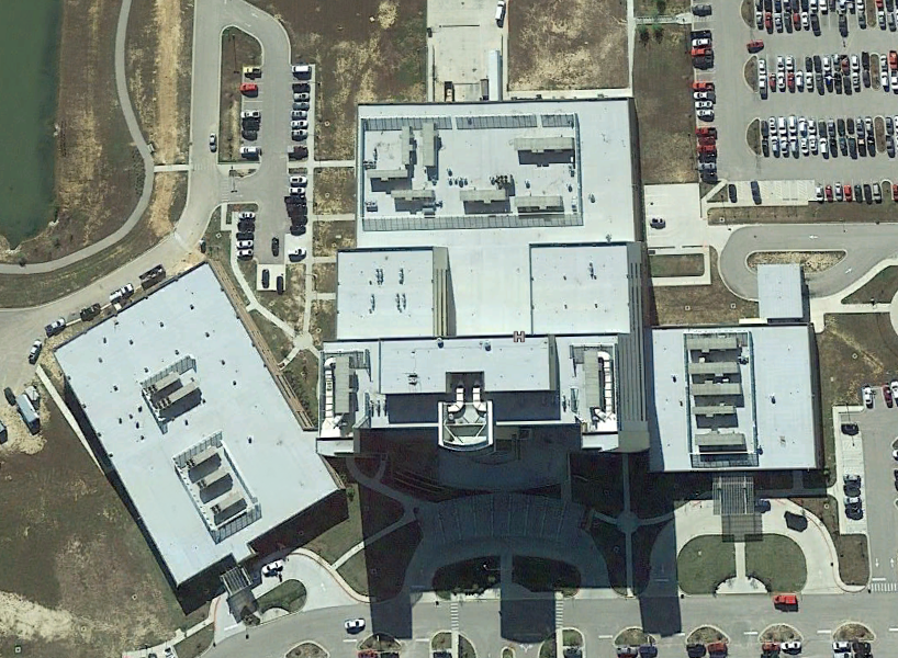 Aerial view of a large, modern building complex with three main rectangular sections features commercial roofing, adjacent parking lots, surrounding roads, and landscaped pathways. Numerous parked cars are visible around the buildings.