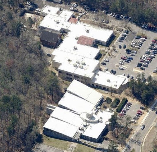 Aerial view of a white-roofed building complex surrounded by trees, parking lots filled with cars, and adjacent roads—ideal for a commercial roofer specializing in commercial roof repair amid wooded and developed land.