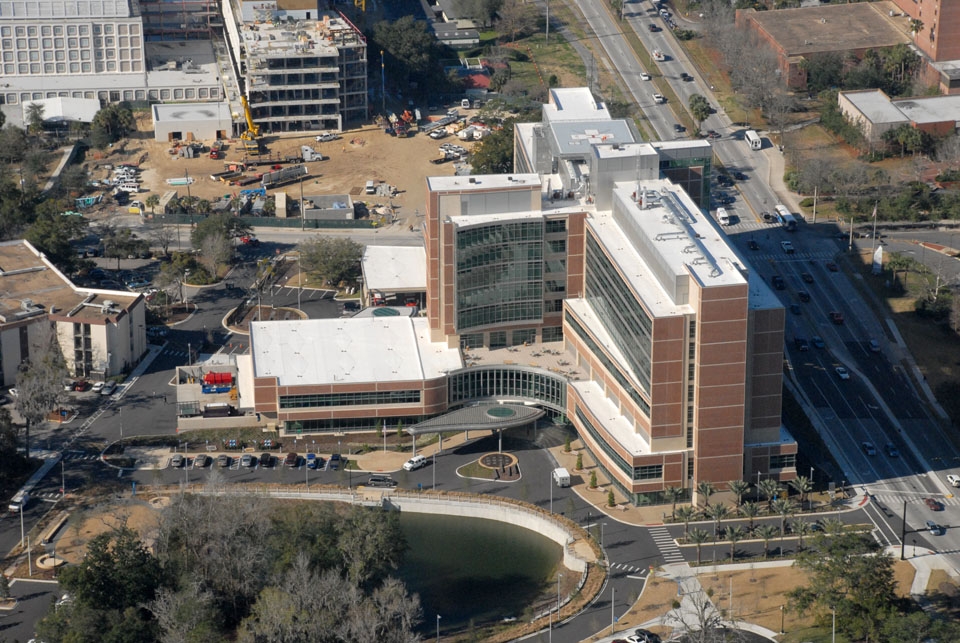Aerial view of a modern multi-story hospital, where a commercial roofer works on the rooftop. Surrounding the building are parking lots, roads, nearby construction sites, and lush trees with a pond in the foreground.