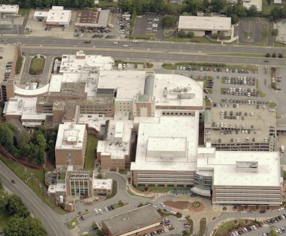 Aerial view of a large hospital complex with multiple connected buildings and white rooftops maintained by commercial roofers, surrounded by parking lots, roads, nearby buildings, green spaces, and trees.