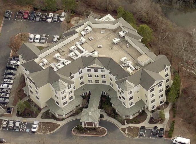Aerial view of a large, multi-story building with a flat central roof and several peaked roofs—ideal for a roofing contractor. The site features a covered entrance, parked cars, trees, and landscaped paths.