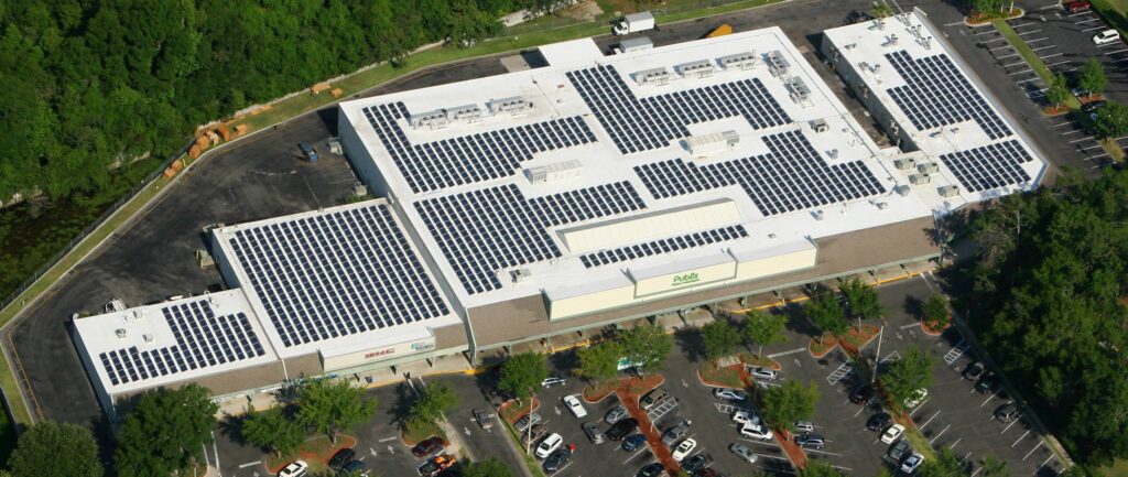 Aerial view of a large supermarket with solar panels covering its white roof, maintained by a professional roofing contractor, surrounded by a parking lot with cars and green trees.