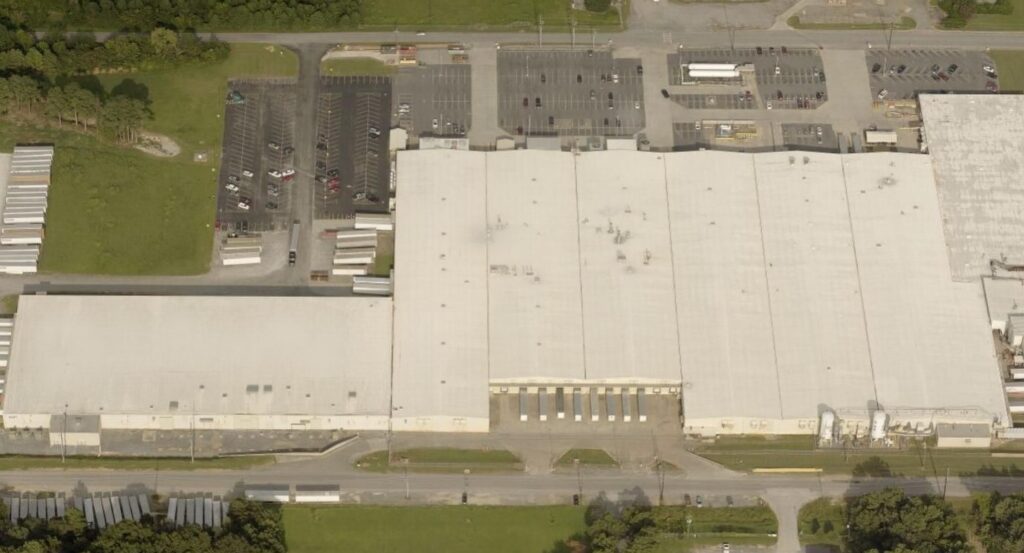Aerial view of a large industrial warehouse with a white roof, multiple loading docks, adjacent parking lots, trailers, and grassy areas—an ideal setting for commercial roofing or expert commercial roof repair services.