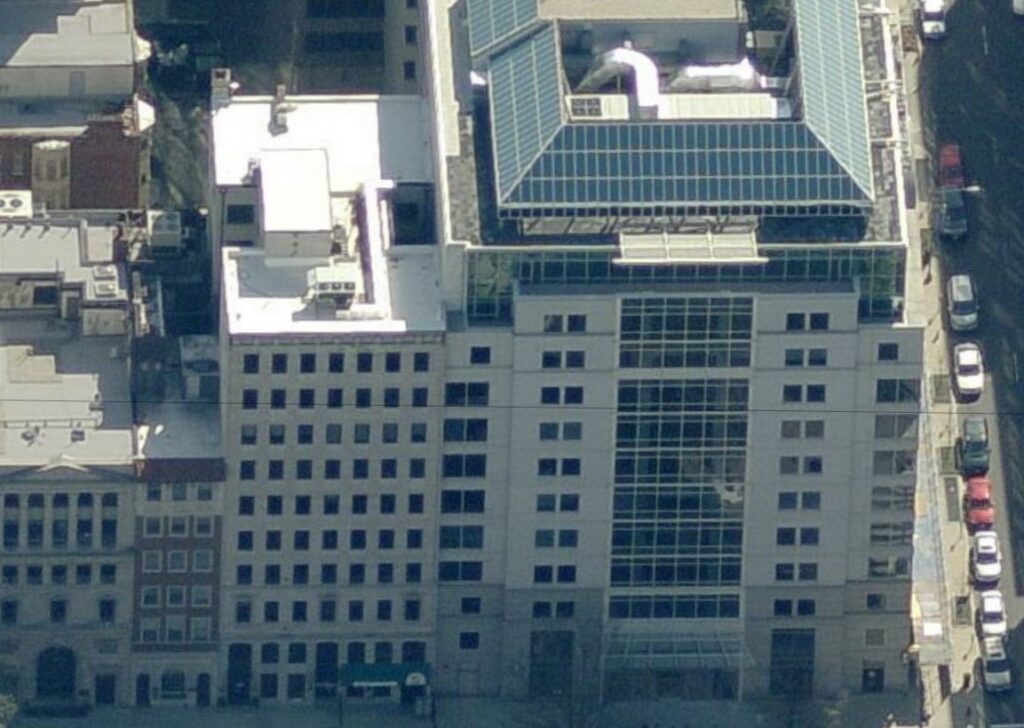 Aerial view of a modern multi-story city building with a glass atrium, solar panels on the roof installed by a commercial roofer, and cars parked along the street below. The building is surrounded by other urban structures.