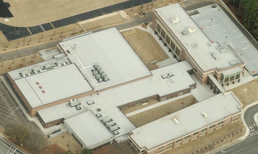 Aerial view of a large, modern school with a flat white roof showcases the quality work of a skilled commercial roofer. Surrounding parking lots, landscaped grounds, and visible main entrance complete the scene.