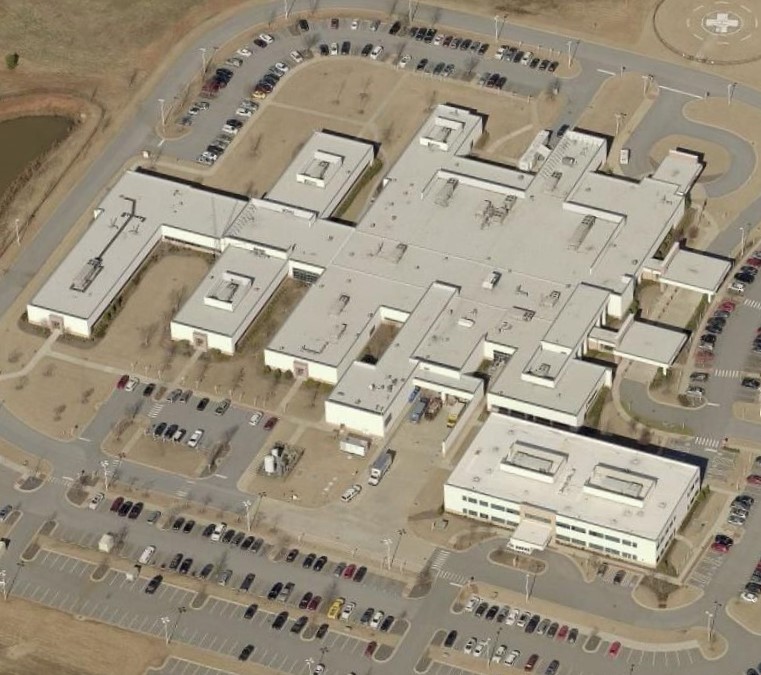 Aerial view of a large, white, single-story building complex with multiple wings—an ideal site for a commercial roofer or roofing contractor—surrounded by several parking lots filled with cars, and bordered by roads and grassy areas.