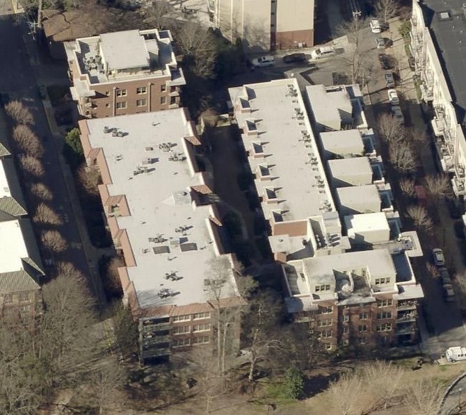 Aerial view of two adjacent rectangular buildings with flat white roofs, possibly serviced by a commercial roofer, surrounded by trees and nearby roads. Shadows from trees and structures are visible on the ground.