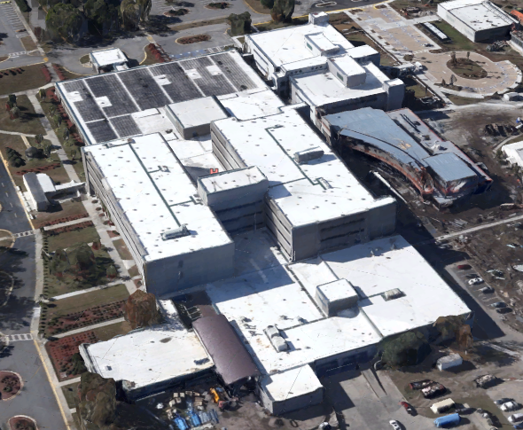 Aerial view of a large, white, flat-roofed building surrounded by parking lots, roads, and landscaping. One side shows ongoing construction with exposed beams and materials as a commercial roofer completes new roofing work.