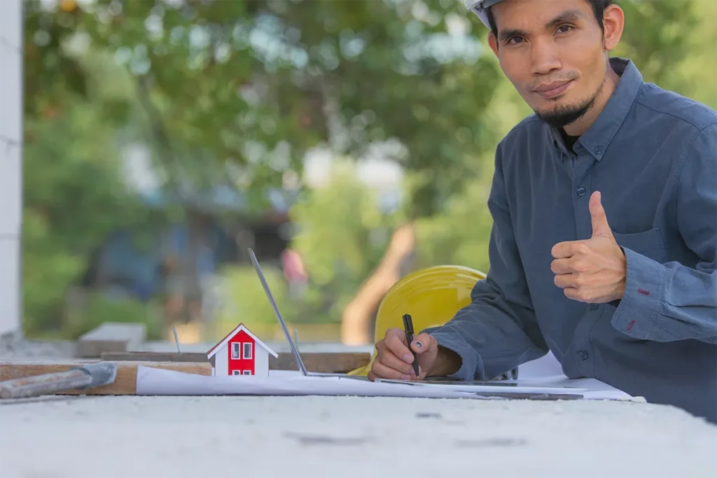 A construction worker in a hard hat gives a thumbs-up while reviewing building plans next to a small house model.