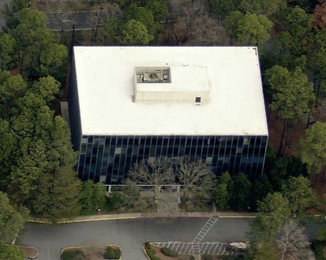 Aerial view of a rectangular office building with a flat roof, surrounded by trees and a parking lot.