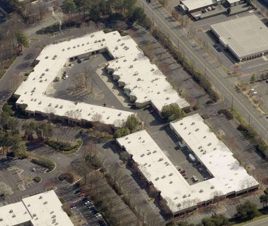 Aerial view of several low-rise commercial buildings with white flat roofs, surrounding parking lots and trees.