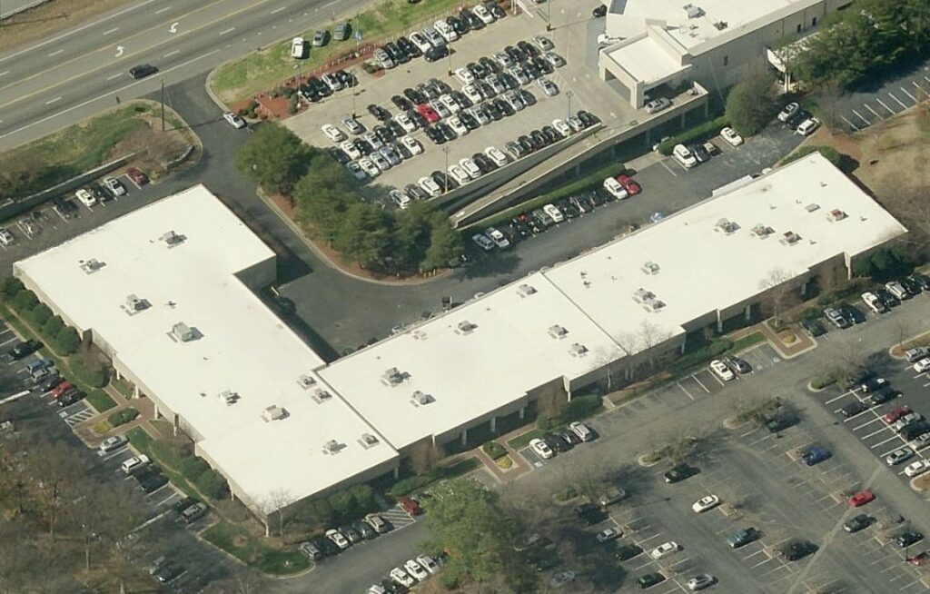 Aerial view of a commercial building complex with flat white roofs and surrounding parking lots filled with cars.