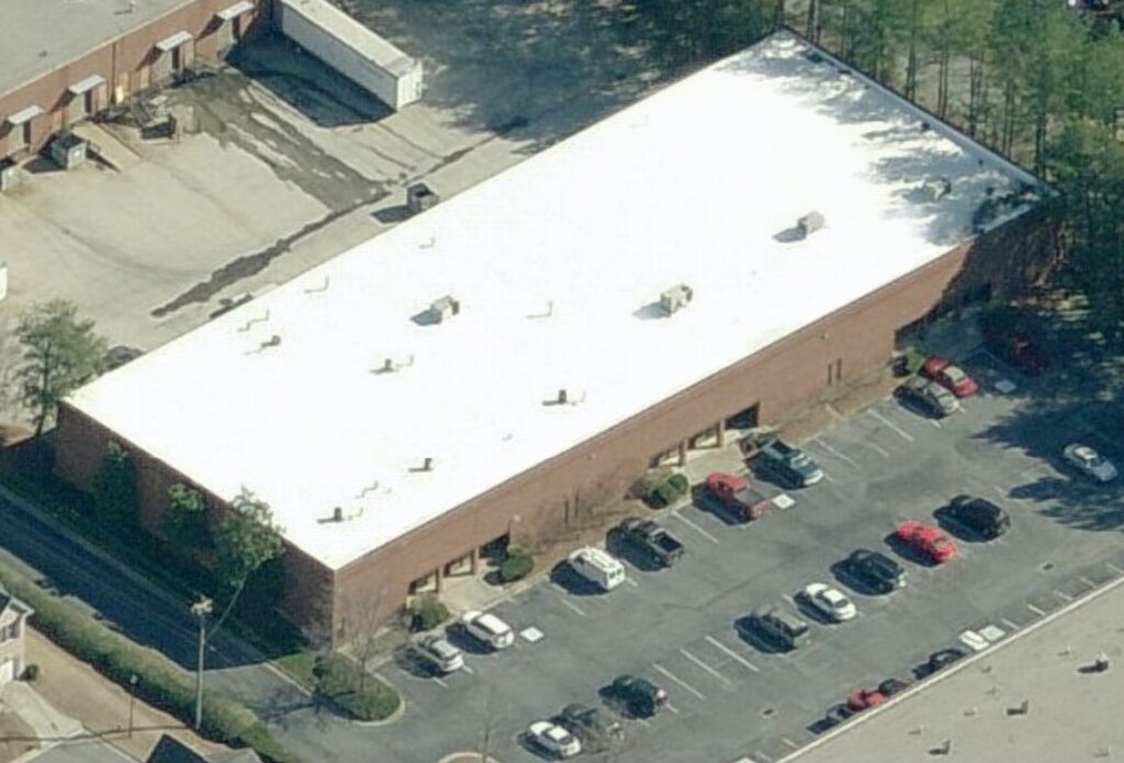 Aerial view of a rectangular brick warehouse with a large white flat roof and an adjacent parking lot filled with cars.