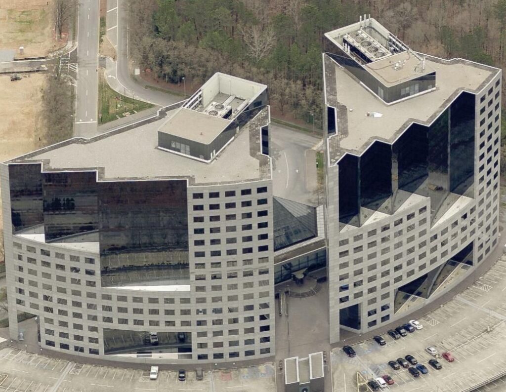 Aerial view of two connected, modern office buildings with geometric shapes and glass facades, surrounded by a parking lot and trees.